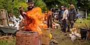 A man tends to a mud furnace with tall flames rising from the top of it, other look on