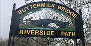 Black metal Buttermilk Bridge Riverside Path sign with writing in yellow and image of a bridge with cow and milk maid crossing