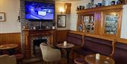 Lounge area of Coast Road Inn with unlit fire in background, with wooden tables. lounge seats and stools plus wall mounted television and cabinet wall