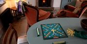 Sitting area in the Barbican. One table with chairs. A game of scrabble is shown on the table. Two arm chairs and a lit fireplace are shown.