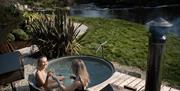 Two people sitting in a small outdoor hot tub beside a river, surrounded by greenery and landscaped gardens.