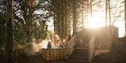 A couple perched on the edge of a hot tub elevated above the Forest floor. Views behind them of the River Maine.