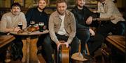 Five men are seated in a cozy pub, smiling at the camera. They have pints of Guinness, a violin, and a drum, creating a warm, musical atmosphere.