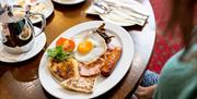 Plate of cooked breakfast with fried egg, sausage, bacon, toast, tomato, and mushrooms on a table with cutlery and a coffee pot.