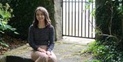 A woman with brown hair is smiling at the camera. She is sitting outside, by a stone wall with an iron gate and is shaded by leafy trees