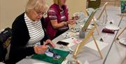 women sat in Midtown Makers painting on small canvas