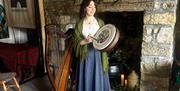 A musician stands beside a harp in front of a stone fireplace, holding a traditional Irish bódhran  in a rustic room lit by candles