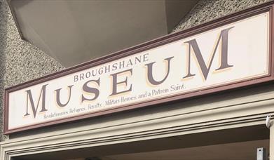 Red and Cream sign on building stating Broughshane Museum - Revolutionaries, Refugees, Royalty, Military Heroes and a Patron Saint.