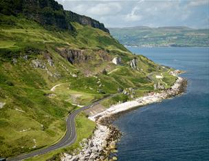 The Causeway Coastal Route running along the side of the ocean with green hills to the other side.