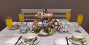 Afternoon tea table setting with 3 tier stand of food plus cups and saucers and bubbly drink.