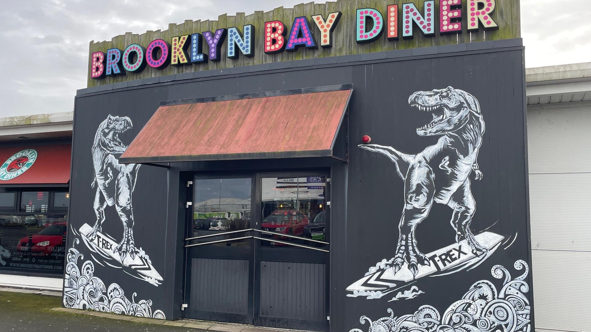 Exterior of Brooklyn Bay Diner entrance with large sign, door canopy and dinosaur wall murals