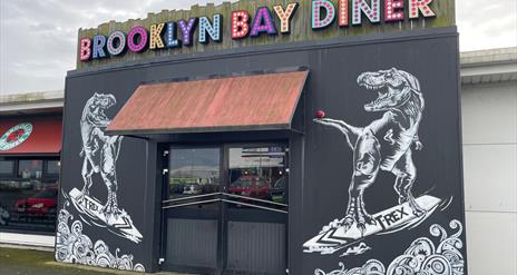 Exterior of Brooklyn Bay Diner entrance with large sign, door canopy and dinosaur wall murals