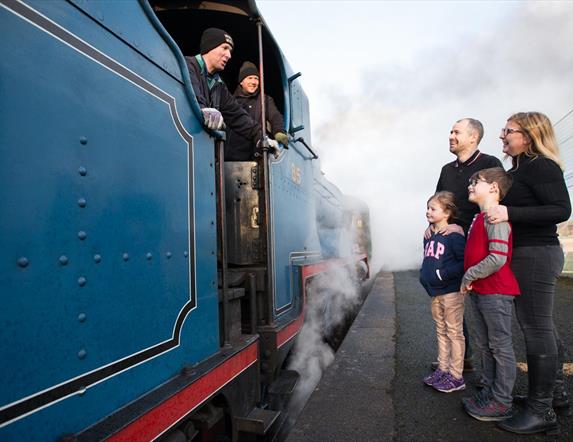 A family of four standing on the platform talking to the driver of a blue steam train
