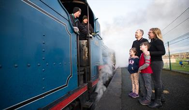 A family of four standing on the platform talking to the driver of a blue steam train