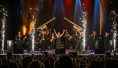 Female singer stood in the middle of brass band on a lit up stage