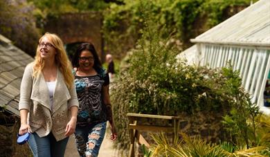 Two women walking through the formal gardens at Greenway. The image is framed by blooming flowers beds, and a glimpse of the wooden framed greenhouse