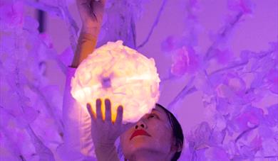 A women in white stands before a magic tree holding a ball of light, there is a wash of purple light