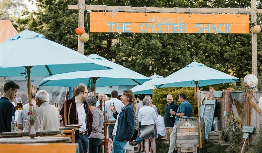 A busy restaurant with customers gathering under a wooden sign reading "Welcome to The Oyster Shack." People are socializing under teal umbrellas, hol