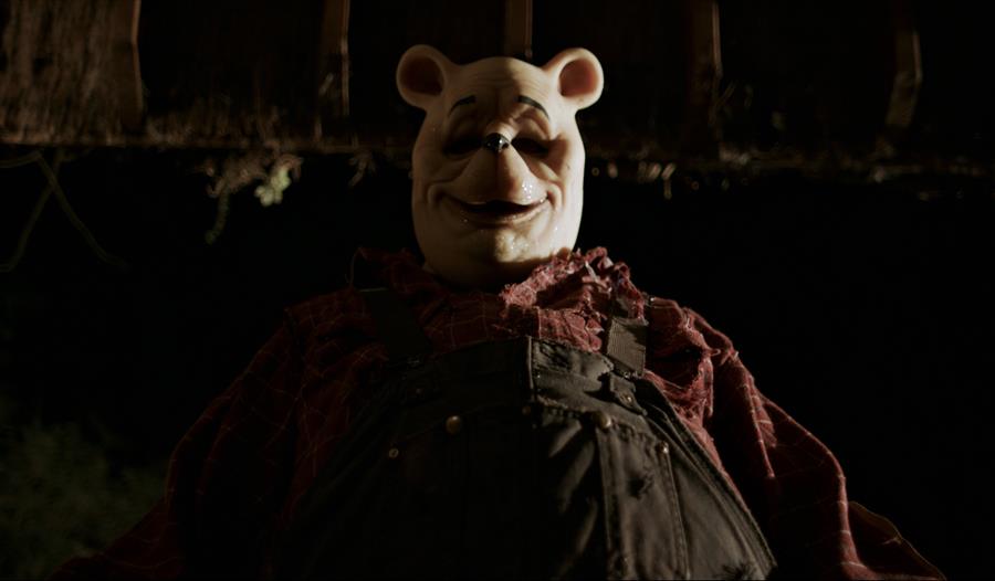 A large person wearing black overalls, a red shirt and a bear mask stands against a dark blurry background