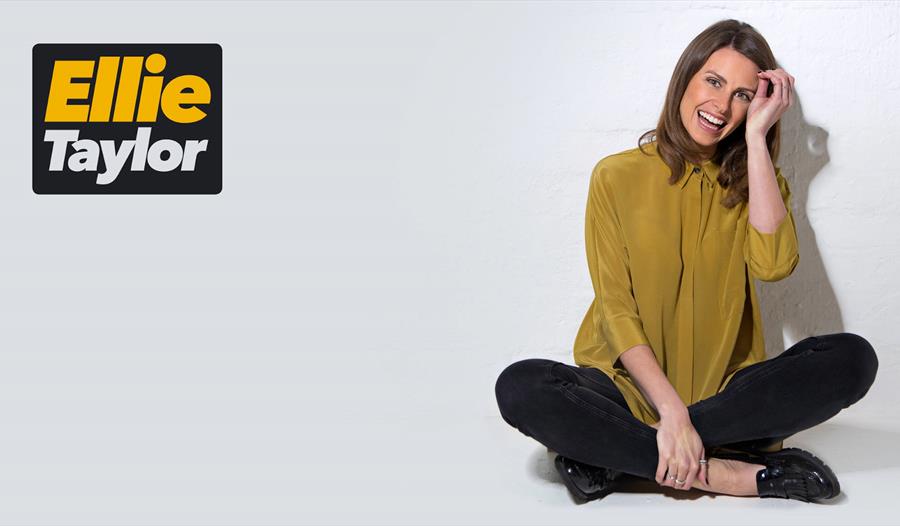 Ellie Taylor sat on a floor with her legs crossed. She is smiling with one hand wiping the hair out of her face.