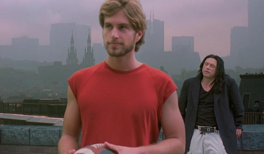 A man in a red shirt holding an American football stands with his back to another man with long black hair. Behind them is a city skyline