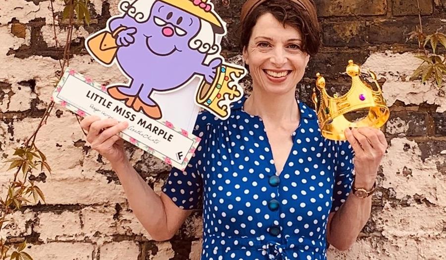 Storyteller in a blue dress, holding a cut out of Little Miss Marple and a crown