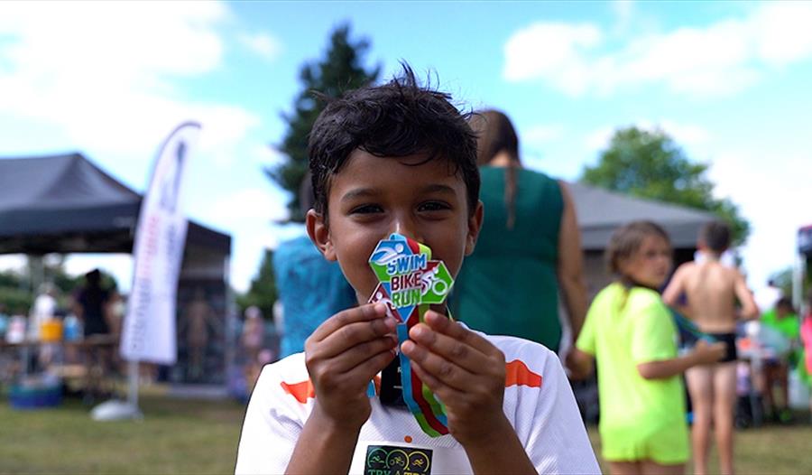 Young boy holding up swim bike run medal in front of face, smiling