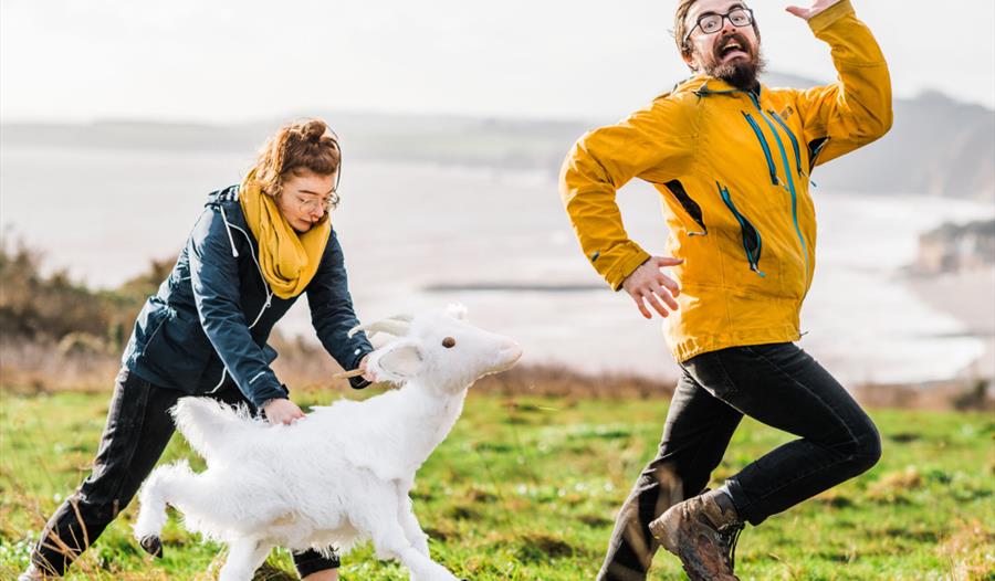 A woman puppeteers a white goat chasing a man in a field