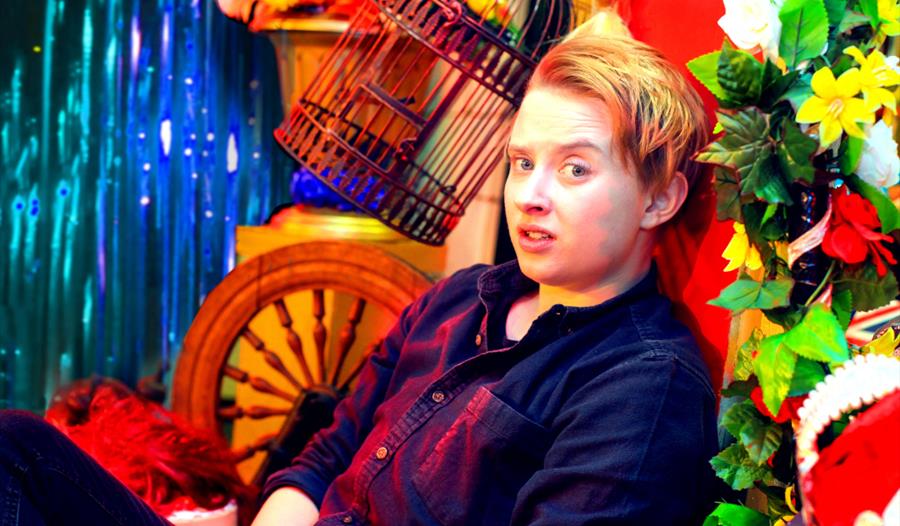 A woman sits amongst a colourful set including flowers, a bird cage, a wooden wheel and tinsel