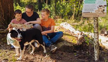 A family and their collie sitting in the forest in front of a Forest Walkies activity panel