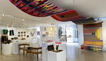 Wide shot of a gallery space showing a range of contemporary craft, including a very large colourful textiles weaving suspended from the ceiling, a be