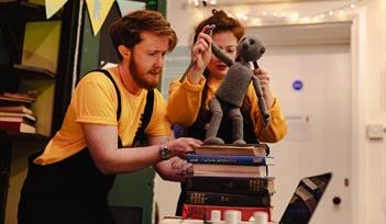 Two people with dark red hair wearing mustard yellow tops and black dungarees, puppeteer a puppet on top of a stack of books