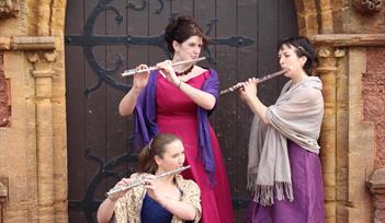 Three women in bright ballroom gowns stand in front of an old wooden church-like door, each holding a flute