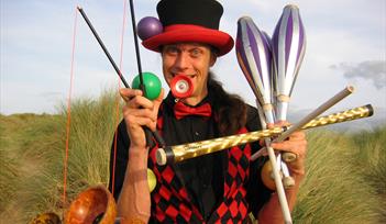 A circus performer holding lots of equipment in a comedic way including juggling batons and diablos.