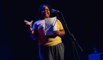 A black woman in a yellow t-shirt holds a script as she talks into a microphone