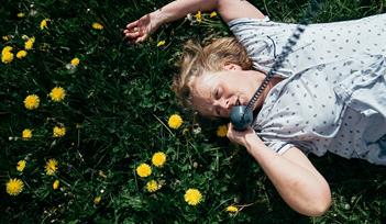 A older white women with blonde hair wearing pyjamas laying on grass, there yellow dandelions around her, she is talking into a corded telephone.