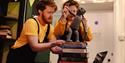 Two people with dark red hair wearing mustard yellow tops and black dungarees, puppeteer a puppet on top of a stack of books