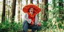 A woman is sitting in a forest wearing a large shiny mushroom hat, and smiling into the camera
