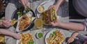 Overhead view of diners sharing fish and chips, sandwiches, salad, and drinks at an outdoor wooden table