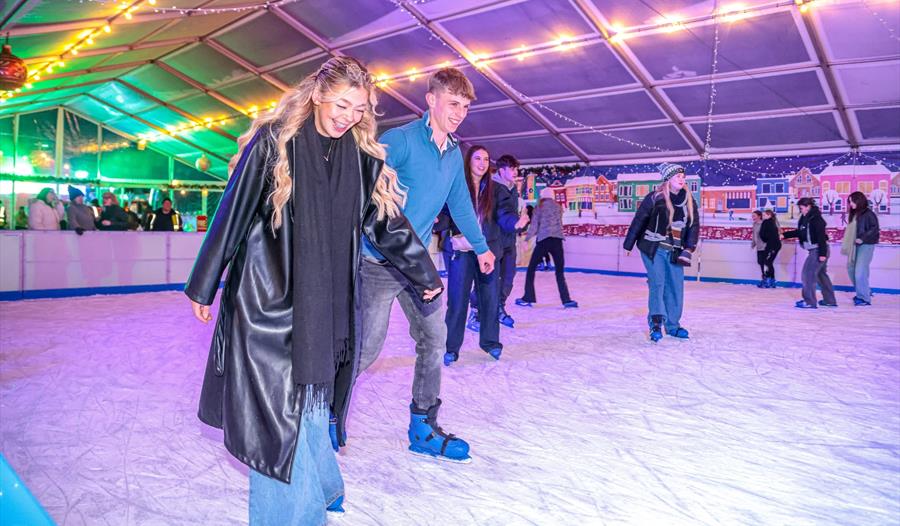 Smiling young couple skating together on the undercover real ice rink at Exeter Winter Wonderland, with other skaters behind them, fairy lights strung Smiling young couple skating together on the undercover real ice rink at Exeter Winter Wonderland, with other skaters behind them, fairy lights strung
