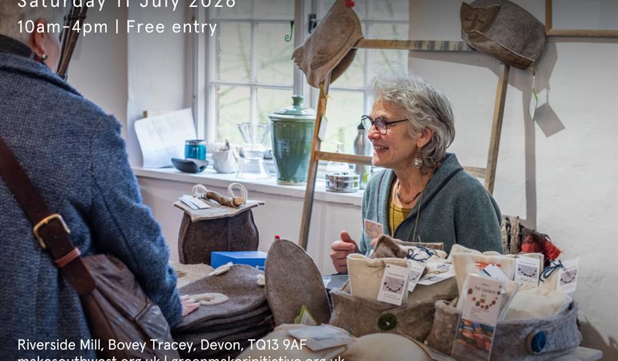Woman with greying hair and glasses sits behind a stall filled with handmade felt items at a craft fair. A woman in a coat and cross body bag is stood Woman with greying hair and glasses sits behind a stall filled with handmade felt items at a craft fair. A woman in a coat and cross body bag is stood