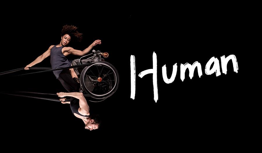 Black background. A photo of a woman and a man swinging in the air like a pendulum, looking powerful and graceful. They share a wheelchair which is he