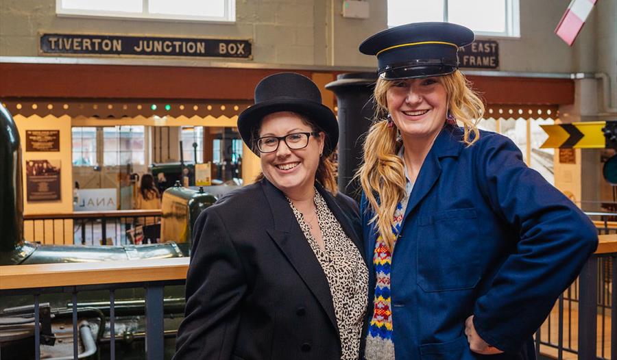 Two museum visitors in historical railway-inspired outfits pose beside a steam engine in Tiverton Museum's transport gallery. Two museum visitors in historical railway-inspired outfits pose beside a steam engine in Tiverton Museum's transport gallery.