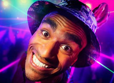 A young man is grinning intensely at the camera. He is wearing a bucket hat with donkey ears. The background is a colourful rave with purple and green