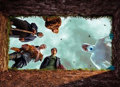 A view from inside a grave looking up at five people gathered around the opening. The group is an eclectic mix: a man in a hazmat suit and mask, a man