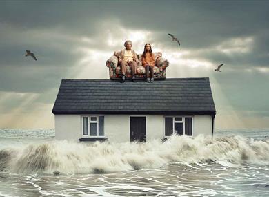 Grey ocean waves crash against the sides of a home. On the roof of the house, is a floral sofa with a young girl and her grandmother sat on, they both