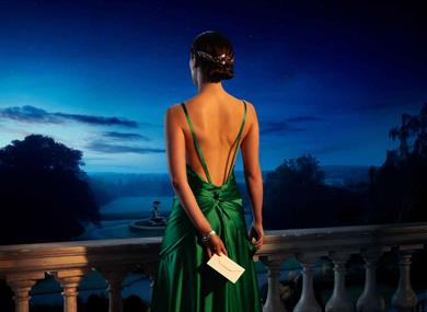 A young woman in a striking green backless dress holds a white envelope in one hand behind her back. She looks out at a garden and fountain from a bal