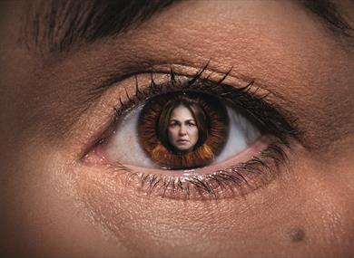 A close up of a woman's eye, within the pupil is an image of the same woman. She has dark brown hair and has a serious expression on her face.