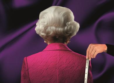 An imitation of Queen Elizabeth II from behind: an older woman with nearly styled, short, grey, wavy hair, pearl earrings and a pink jacket. A hand ho