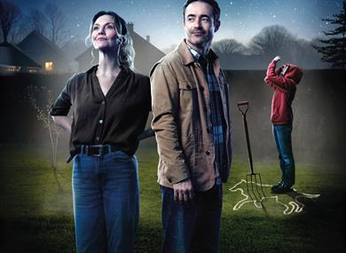 A casually dressed woman and man stand in a garden at night-time, against silhouetted houses, trees and a starry sky. Behind them, a young person in a